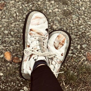 May include: A pair of white slip-on sneakers featuring a brown and tan feather print pattern. The shoes have a thick black rubber sole, a textured grey fabric trim around the base, and cream-colored laces tied in a bow at the center front.