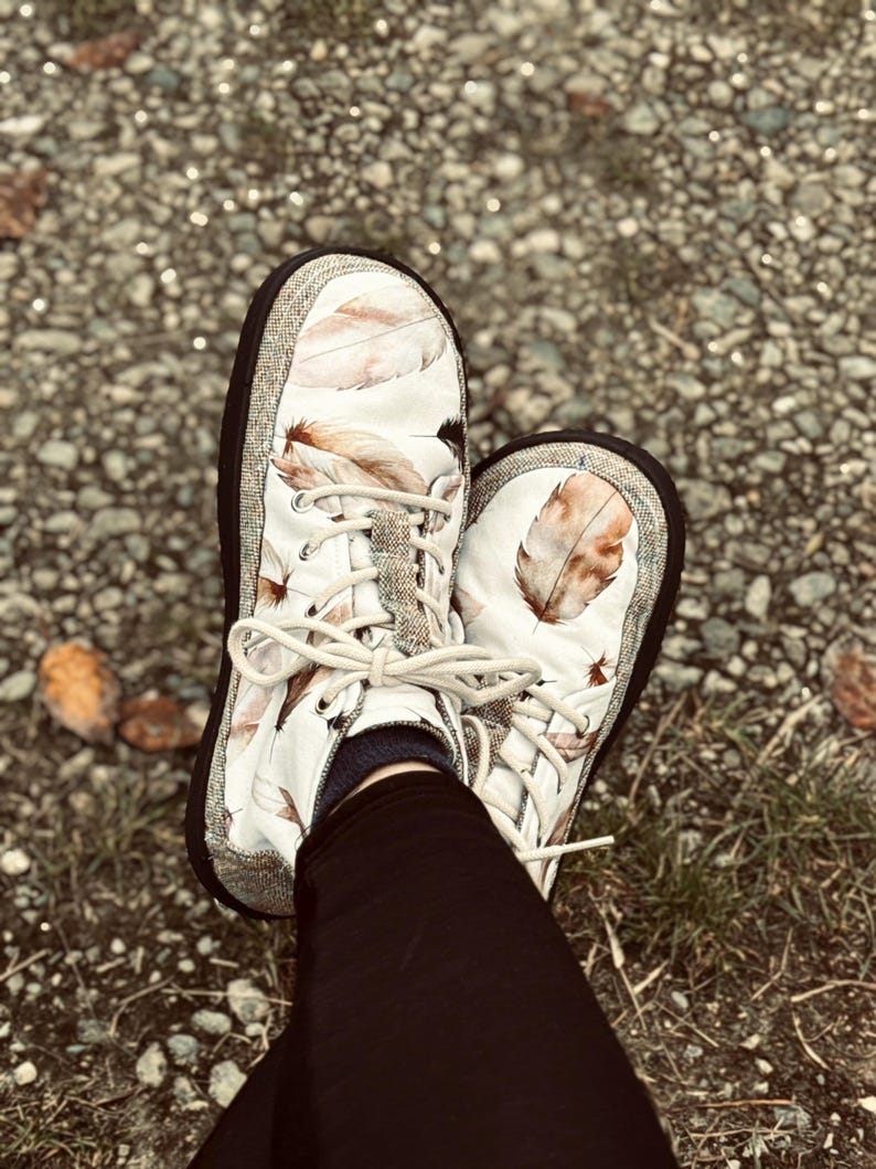 May include: A pair of white slip-on sneakers featuring a brown and tan feather print pattern. The shoes have a thick black rubber sole, a textured grey fabric trim around the base, and cream-colored laces tied in a bow at the center front.