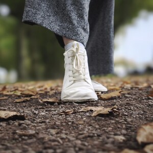 May include: A white lace-up sneaker with a rounded toe and a flexible sole, paired with grey herringbone patterned trousers. The footwear features a simple design with matching white laces and a low-profile silhouette, shown in a neutral color.