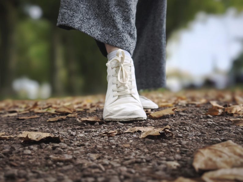 May include: A white lace-up sneaker with a rounded toe and a flexible sole, paired with grey herringbone patterned trousers. The footwear features a simple design with matching white laces and a low-profile silhouette, shown in a neutral color.