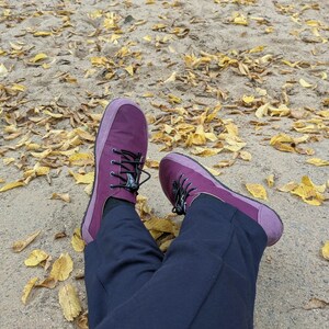 May include: A pair of deep purple sneakers with black laces and a rounded toe box, worn with dark navy blue trousers. The footwear features a solid purple upper with a slightly lighter purple trim around the sole and a thin black rubber outsole.
