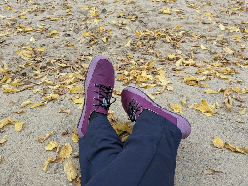May include: A pair of deep purple sneakers with black laces and a rounded toe box, worn with dark navy blue trousers. The footwear features a solid purple upper with a slightly lighter purple trim around the sole and a thin black rubber outsole.