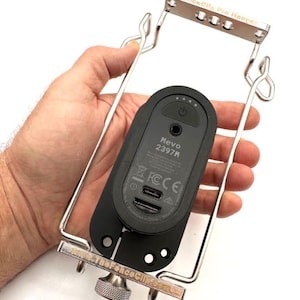 May include: A silver metal phone holder with a black phone attached. The phone holder has a knob on the bottom and the text "TheForceClip.com" is engraved on the side. The phone has the text "Mevo 2397M" and "FCC CE" on the back.