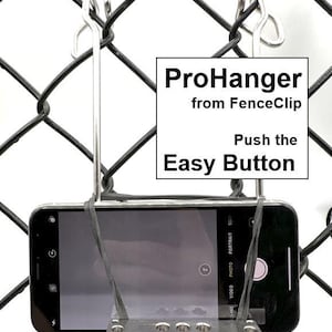 May include: A silver ProHanger from FenceClip attached to a black chain link fence. A smartphone is secured to the hanger with a black rubber band. The text on the hanger reads "ProHanger from FenceClip Push the Easy Button" and "www.TheFenceClip.com".
