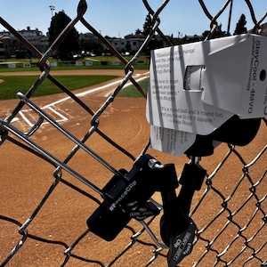 May include: A black #FenceClip attached to a chain-link fence, holding a white sign with text about baseball. The background shows a baseball field with a dirt infield and green outfield.