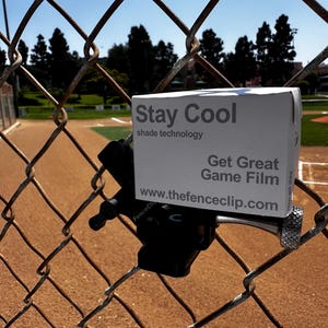 May include: A white box with the text "Stay Cool shade technology" and "Get Great Game Film" is attached to a chain-link fence. The website address www.thefenceclip.com is also visible. The background shows a baseball field.