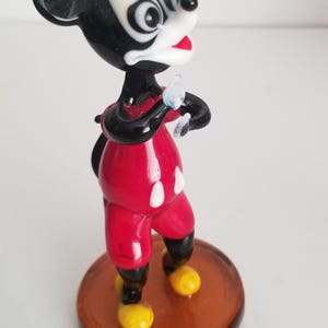 Puede incluir: Una figurita de vidrio de Mickey Mouse con su caracter&iacute;stico pantal&oacute;n rojo y zapatos amarillos. Est&aacute; de pie sobre una base marr&oacute;n y tiene una cara en blanco y negro.