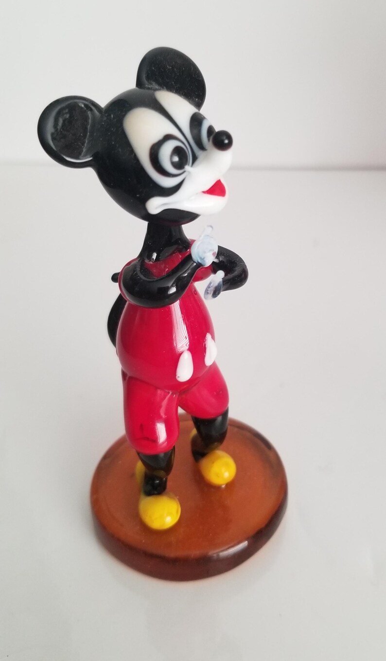 Puede incluir: Una figurita de vidrio de Mickey Mouse con su caracter&iacute;stico pantal&oacute;n rojo y zapatos amarillos. Est&aacute; de pie sobre una base marr&oacute;n y tiene una cara en blanco y negro.