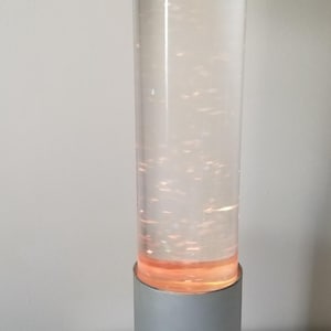 Könnte beinhalten: Eine silberne und transparente Lavalampe mit zylindrischem Design. Die Lampe hat einen silbernen Sockel und eine silberne Oberseite, mit einem transparenten Glasrohr, das eine wirbelnde, orangefarbene Flüssigkeit enthält. Die Lampe steht auf einem schwarzen, kreisförmigen Sockel.