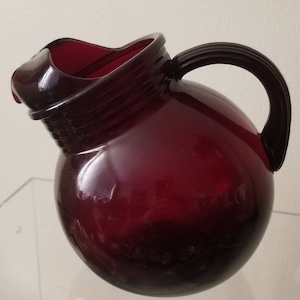 May include: A deep red glass pitcher with a round body and a handle. The pitcher has a textured rim and a smooth, glossy finish.