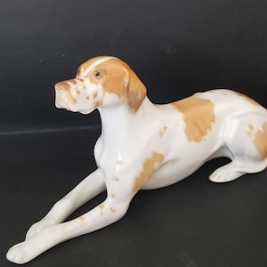 Puede incluir: Figura de porcelana de un perro en posición de descanso. El perro tiene un pelaje blanco con manchas marrones en la cabeza, las orejas y el cuerpo. La figura es detallada y parece ser de raza Pointer. El perro está tumbado con las patas delanteras extendidas.