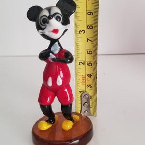 Puede incluir: Una figurita de vidrio de Mickey Mouse de pie con los brazos cruzados. Lleva pantalones cortos rojos con c&iacute;rculos blancos, zapatos negros y una cara en blanco y negro. La figurita mide aproximadamente 10 cm de alto y est&aacute; de pie sobre una base marr&oacute;n.