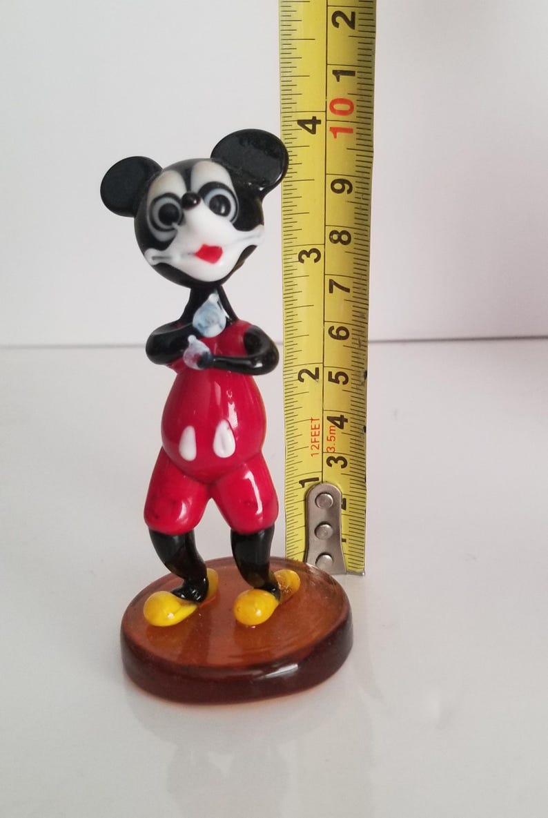 Puede incluir: Una figurita de vidrio de Mickey Mouse de pie con los brazos cruzados. Lleva pantalones cortos rojos con c&iacute;rculos blancos, zapatos negros y una cara en blanco y negro. La figurita mide aproximadamente 10 cm de alto y est&aacute; de pie sobre una base marr&oacute;n.