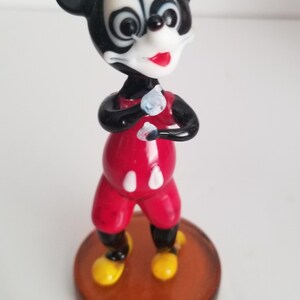 Puede incluir: Una figurita de vidrio de Mickey Mouse con un atuendo rojo y blanco, de pie sobre una base marr&oacute;n. La figurita tiene orejas negras, una nariz negra y ojos blancos.