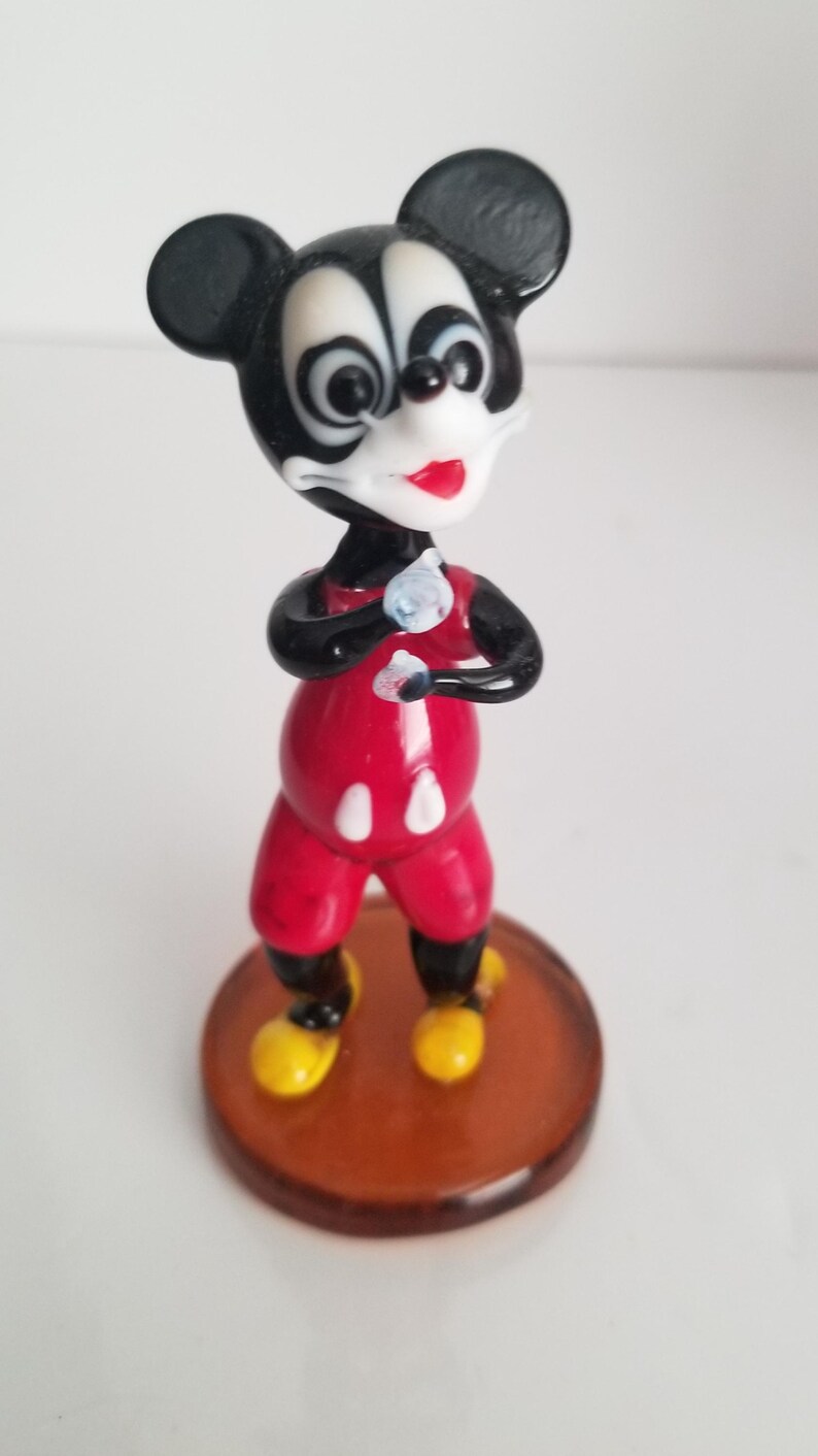 Puede incluir: Una figurita de vidrio de Mickey Mouse con un atuendo rojo y blanco, de pie sobre una base marr&oacute;n. La figurita tiene orejas negras, una nariz negra y ojos blancos.