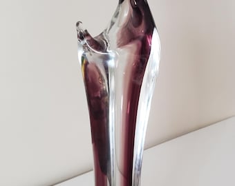 Puede incluir: Una escultura de vidrio alta y esbelta de una figura estilizada con la cabeza inclinada y un halo. La pieza está hecha de vidrio transparente con remolinos de color púrpura intenso integrados en el centro, sobre una base circular.