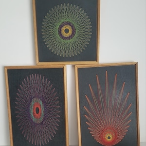 May include: Three framed string art pieces. Each features a geometric design with colorful threads on a dark background. The frames are made of light-colored wood. The designs include circular and radial patterns.