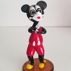 Puede incluir: Una peque&ntilde;a figura de vidrio de Mickey Mouse. La figura es negra y roja con zapatos amarillos. Mickey est&aacute; de pie con los brazos cruzados y tiene una cara blanca con ojos negros y una boca roja.