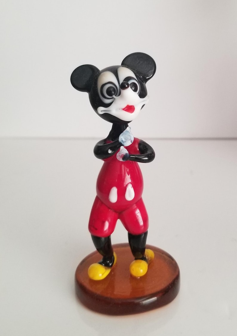 Puede incluir: Una peque&ntilde;a figura de vidrio de Mickey Mouse. La figura es negra y roja con zapatos amarillos. Mickey est&aacute; de pie con los brazos cruzados y tiene una cara blanca con ojos negros y una boca roja.
