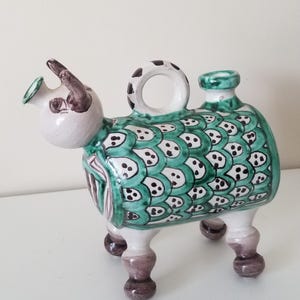 May include: A ceramic figurine in the shape of a stylized animal, featuring a white head with brown horns and a green body with a scale-like pattern. The figurine has a handle and a spout, and stands on four legs with brown feet.