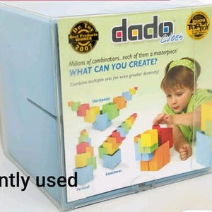 Puede incluir: Una caja de plástico transparente que contiene un juego de juguetes "dada Cubes". La caja muestra a un niño jugando con bloques de colores. El texto en la caja incluye "Best Products Winner 2001" y "WHAT CAN YOU CREATE?" También se ven las palabras "gently used".
