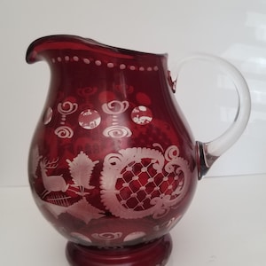Peut inclure: Un pichet en verre rouge avec une anse transparente. Le pichet est décoré de motifs gravés blancs représentant un cerf, des fleurs et des motifs géométriques.