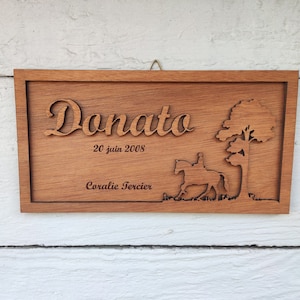 Può includere: Un cartello in legno con il nome "Donato" inciso in corsivo, la data "20 juin 2008" e il nome "Coralie Tercier". Il cartello presenta anche una silhouette di un cavallo e un cavaliere con un albero sullo sfondo.
