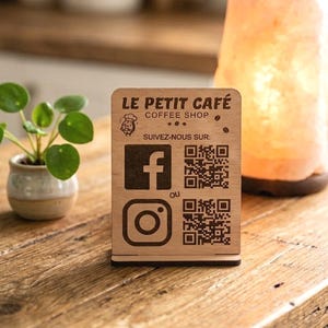 May include: A wooden sign for "Le Petit Café" with Facebook and Instagram logos and QR codes. The sign is on a wooden surface, next to a small plant, a Himalayan salt lamp, and a coffee cup.