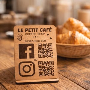 May include: A rectangular wooden sign with a stand featuring the text LE PETIT CAFE COFFEE SHOP, a small chef icon, and the text SUIVEZ-NOUS SUR. It includes Facebook and Instagram logos paired with two QR codes and the word OU in the center.
