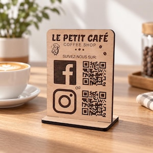 Peut inclure: Un panneau rectangulaire en bois clair avec une gravure marron foncé. Il affiche le texte LE PETIT CAFÉ COFFEE SHOP, une icône de chef, ainsi que les logos Facebook et Instagram accompagnés de QR codes et du mot OU entre les deux.