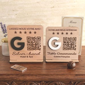 May include: Two wooden signs with the letter "G" and QR codes. One sign says "Silver-hand Hotel & Spa" and the other says "Table Gourmande Cuisine française". A hotel card and a chocolate are on the table.