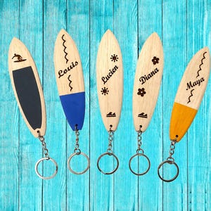 Peut inclure: Cinq porte-clés en forme de planche de surf en bois avec anneaux métalliques. L'un présente un grip noir et une icône de surfeur. Les autres sont personnalisés avec les prénoms Louis, Lucien, Diana et Maya, avec des motifs soleil, fleur et vague.