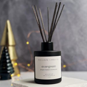 Peut inclure: Diffuseur de roseaux en verre noir avec des roseaux noirs. La bouteille porte une étiquette blanche avec le texte "EVA LOUISE CANDLE CO" et "evergreen" en caractères plus grands. L'étiquette comprend également le texte "juniper | balsam | eucalyptus", "REED DIFFUSER", "NEW GLOUCESTER, ME", et "4 OZ".