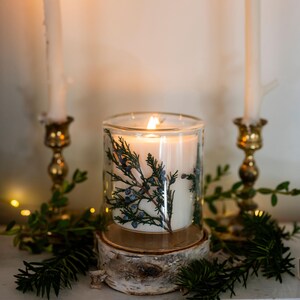 May include: A lit, white candle in a clear glass jar, decorated with sprigs of green and blue berries. The candle sits on a wooden base, surrounded by greenery and flanked by two brass candle holders with lit candles.