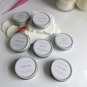 May include: Assortment of six small, round, white tin candle jars with gold lids. Each jar has a different scent label: Coconut Sugar, Woods & Musk, Dark Rose, Blood Orange, Saffron & Tobacco, and Coastal Amber.