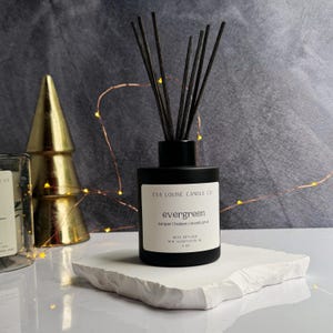 Peut inclure: Diffuseur de roseaux en verre noir avec des roseaux noirs et une étiquette blanche qui indique "EVA LOUISE CANDLE CO" et "evergreen juniper balsam | eucalyptin" avec le texte "REED DIFFUSER NEW GLOUCESTER, NJ 4 OZ" en bas.