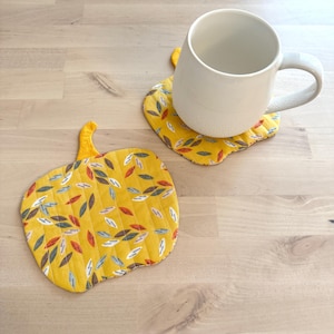 May include: Two yellow pumpkin-shaped quilted fabric coasters with a leaf pattern. One coaster has a white mug on top. The coasters are on a light wood surface. The coasters are a fall-themed home decor item.