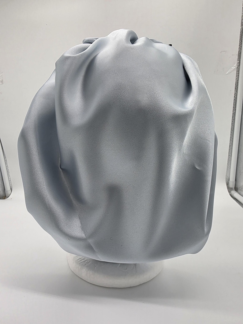 Pu&ograve; includere: Un cappuccio in raso azzurro, progettato per proteggere i capelli. Il cappuccio &egrave; raccolto nella parte superiore e ha una consistenza liscia e lucida. &Egrave; esposto su una forma arrotondata bianca su sfondo bianco.