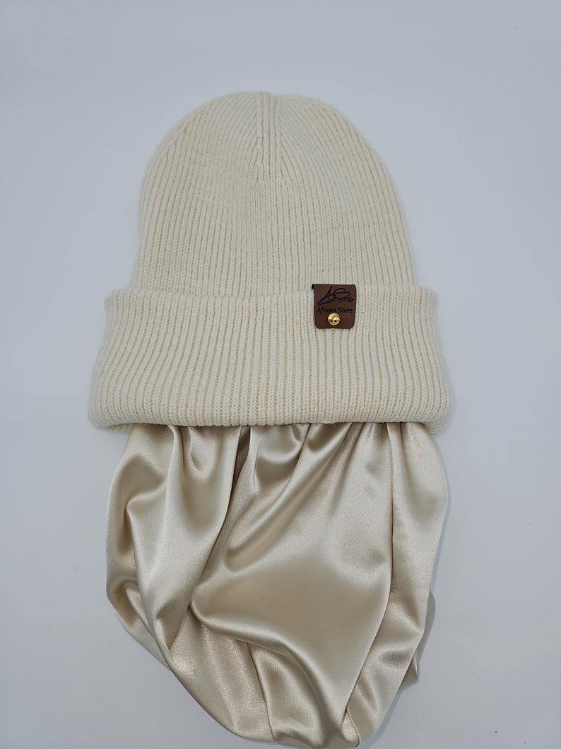 May include: A cream-colored knitted beanie with a brown leather patch that says "House of Yarn" and a gold button. The beanie is sitting on top of a cream-colored satin fabric.