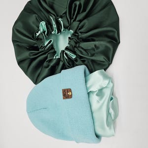 May include: A dark green satin bonnet with a light green interior is positioned above a light blue beanie. The beanie has a small brown leather tag. A light green satin fabric is inside the beanie.