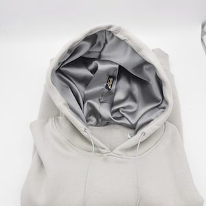 May include: A light gray hooded sweatshirt with a silver satin lining. The hood is folded down and the drawstring is visible.