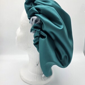 Pu&ograve; includere: Un cappuccio in raso verde acqua con interno bianco, esposto su una testa di manichino bianca. Il cappuccio ha un design arricciato intorno alla testa e una chiusura con coulisse. Il tessuto ha una leggera lucentezza.