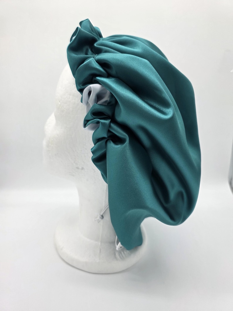 Pu&ograve; includere: Un cappuccio in raso verde acqua con interno bianco, esposto su una testa di manichino bianca. Il cappuccio ha un design arricciato intorno alla testa e una chiusura con coulisse. Il tessuto ha una leggera lucentezza.
