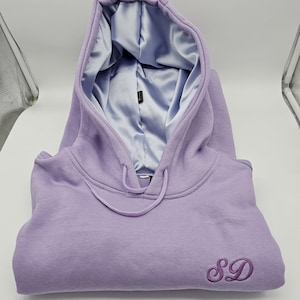 May include: A lavender hoodie with a light blue satin lining in the hood. The hoodie has a drawstring and the embroidered initials "SD" on the front. The garment is folded neatly.
