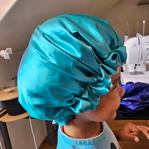 Pu&ograve; includere: Un cappuccio in raso turchese, progettato per proteggere i capelli. Il cappuccio &egrave; raccolto ai bordi e ha un aspetto lucido. La persona che indossa il cappuccio indossa una camicia blu con una grafica a cartoni animati.