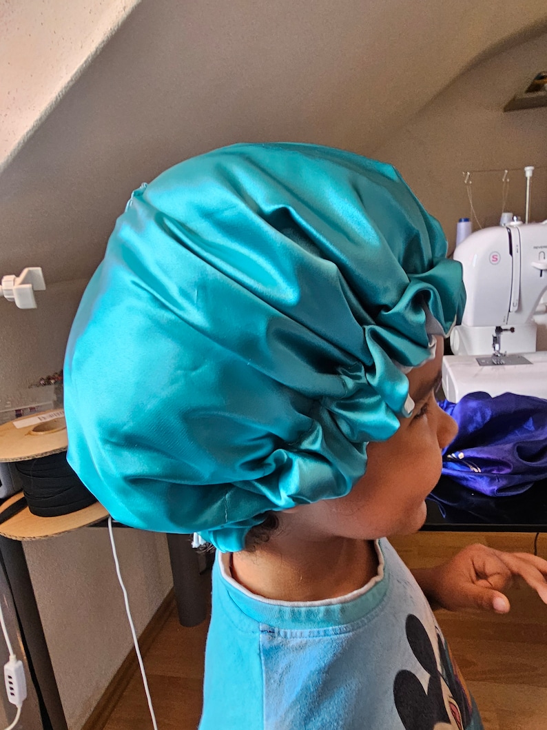 Pu&ograve; includere: Un cappuccio in raso turchese, progettato per proteggere i capelli. Il cappuccio &egrave; raccolto ai bordi e ha un aspetto lucido. La persona che indossa il cappuccio indossa una camicia blu con una grafica a cartoni animati.