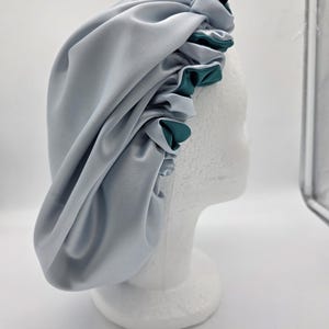 Pu&ograve; includere: Un cappuccio in raso azzurro con finiture verde acqua, esposto su una testa di manichino bianca. Il cappuccio &egrave; raccolto nella parte superiore e ha una consistenza liscia e lucida. &Egrave; visibile una piccola etichetta nera.
