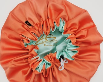Può includere: Un cappello in raso corallo con fodera in raso blu chiaro. Il cappello ha una chiusura con coulisse e il testo "AS Bonnets" è stampato sulla fodera.