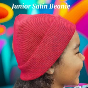 Peut inclure: Un bonnet en satin rouge, avec le texte "Junior Satin Beanie" en blanc, est présenté sur la tête d'une personne. Le bonnet est fait d'une matière douce et côtelée. Le fond est un motif abstrait coloré.