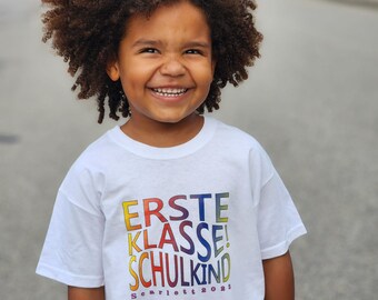 Puede incluir: Una persona joven con una camiseta blanca con las palabras "Erste Klasse! Schulkind" en colores arcoíris y el texto "Scarlett 2023" en negro debajo. La persona sonríe y tiene el pelo rizado castaño.